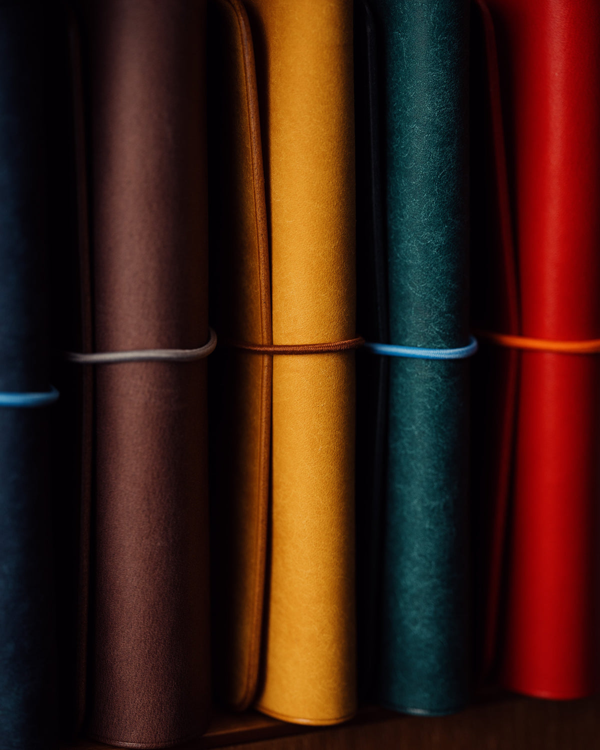 Colourful leather notebook spines and elastic cords side by side in warm light and deep shadow.