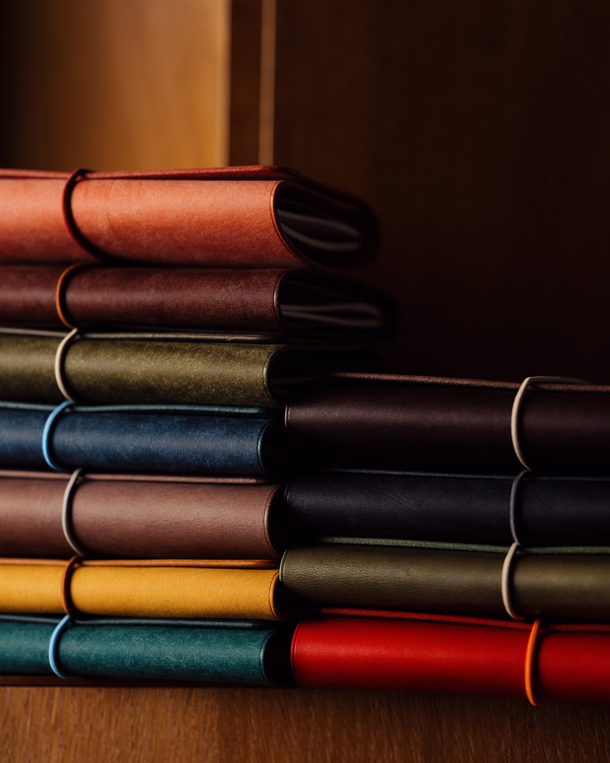 Colourful leather-covered notebooks layered horizontally on a wooden shelf in warm side light.