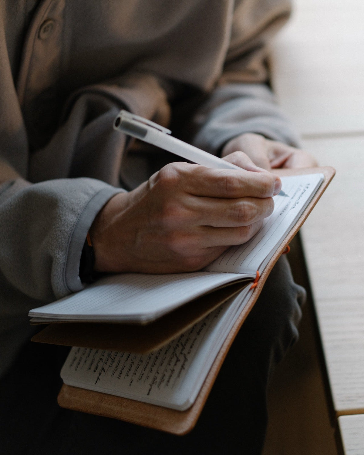 A person writing in an open notebook on their lap, with filled pages resting underneath.