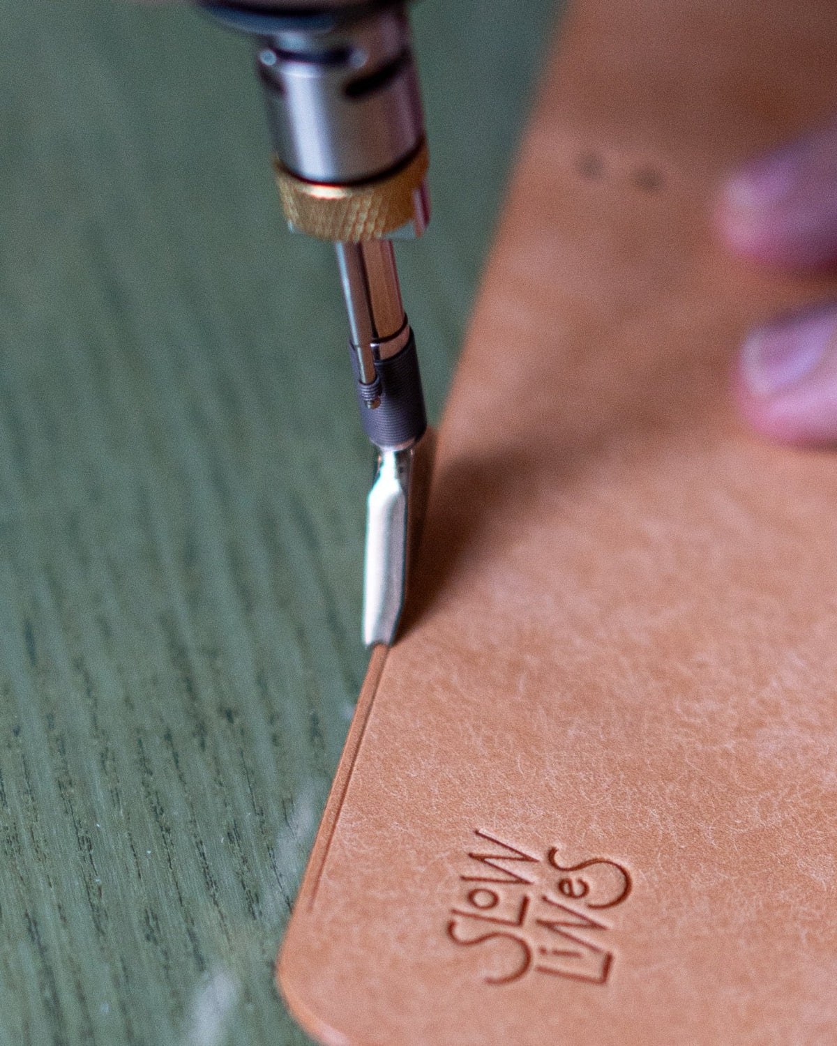 A hand guiding a leatherworking tool along a tan leather cover beside the embossed SLOWLINES mark.