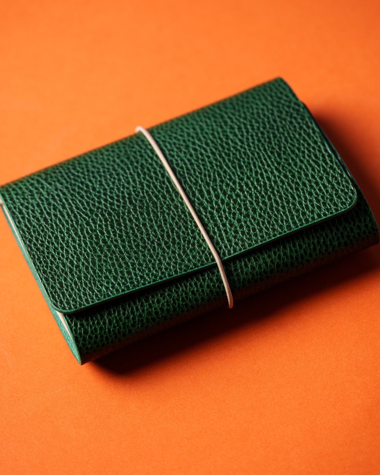 Green pebbled leather trifold cover closed with a pale elastic, on an orange background