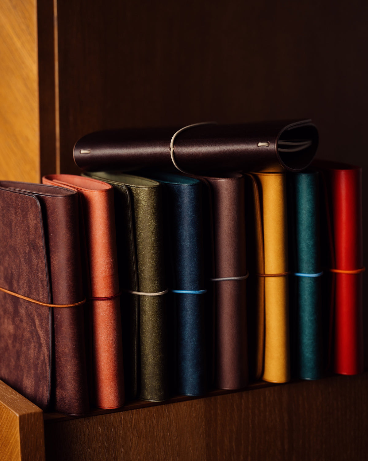 Colourful leather notebooks arranged on a wooden shelf, with one rolled cover resting across the top.